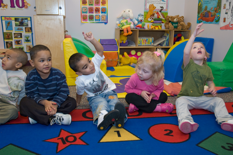 Students in a preschool classroom sitting on a mat and raising their hands.