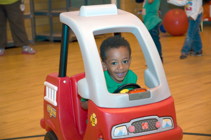 Child sitting in a Cozy Coop fire truck in a gymnasium setting.