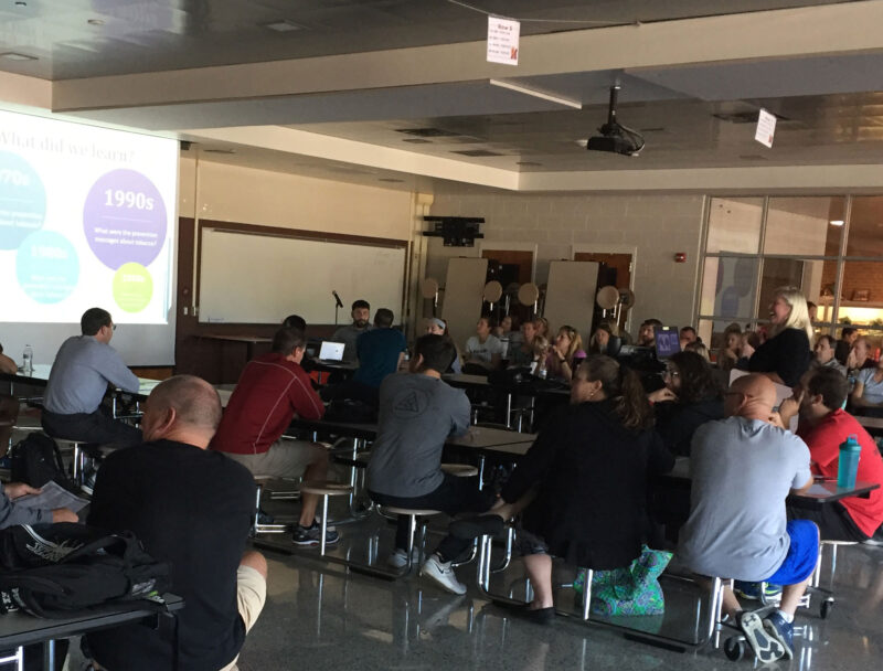 Photo of a school cafeteria filled with educators for a professional development event. A powerpoint presentation can be seen on the screen.