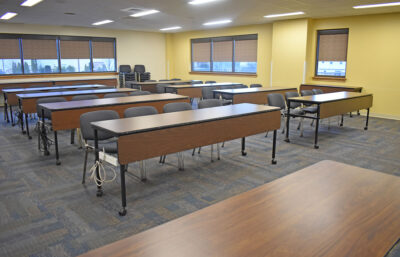 Photo of a yellow-painted room set up with four rows of a wooden tables in a lecture hall style with four gray chairs at each table.