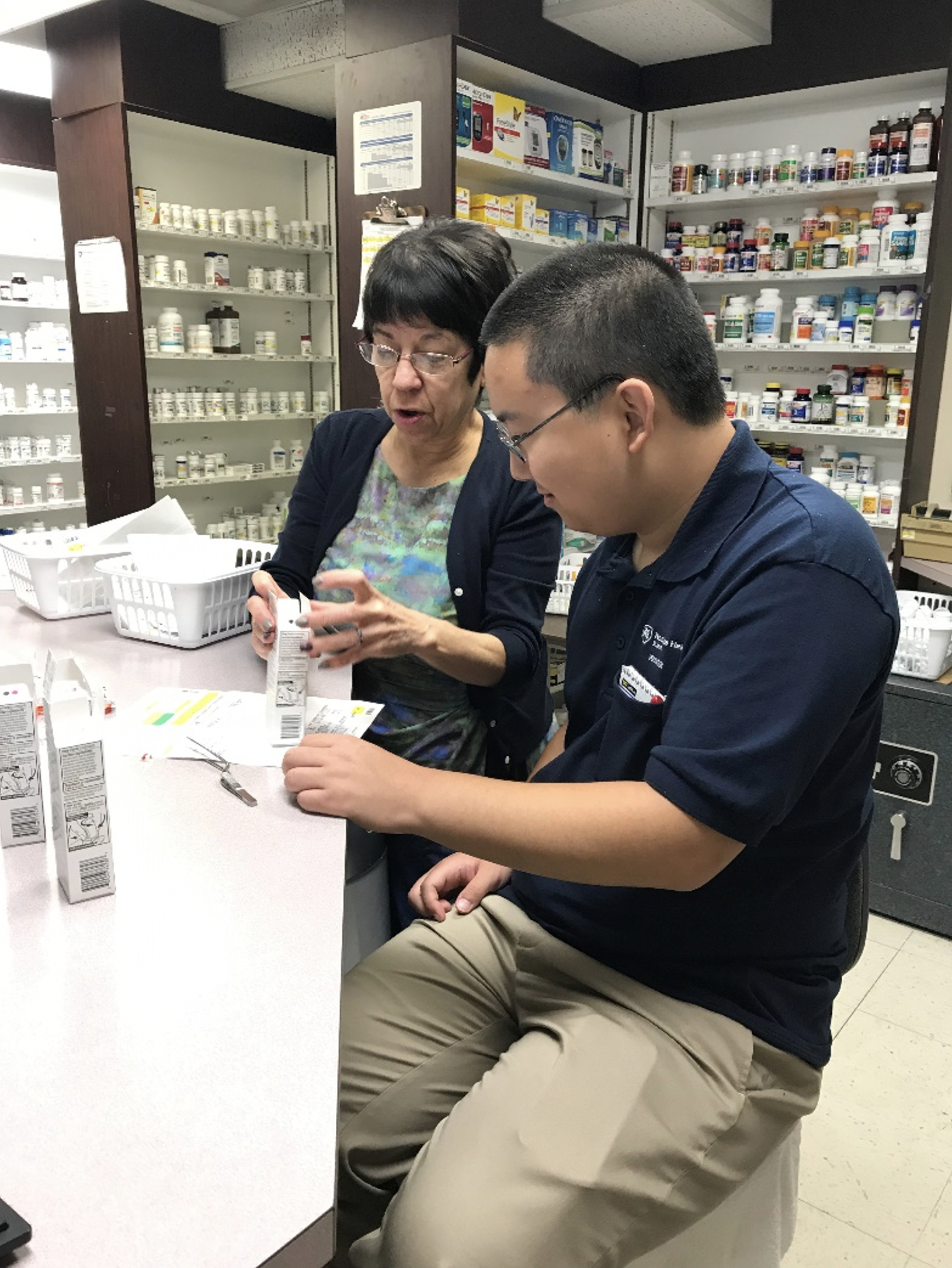 A Project SEARCH intern and his supervisor sit at a pharmacy counter as she shows him a prescription box