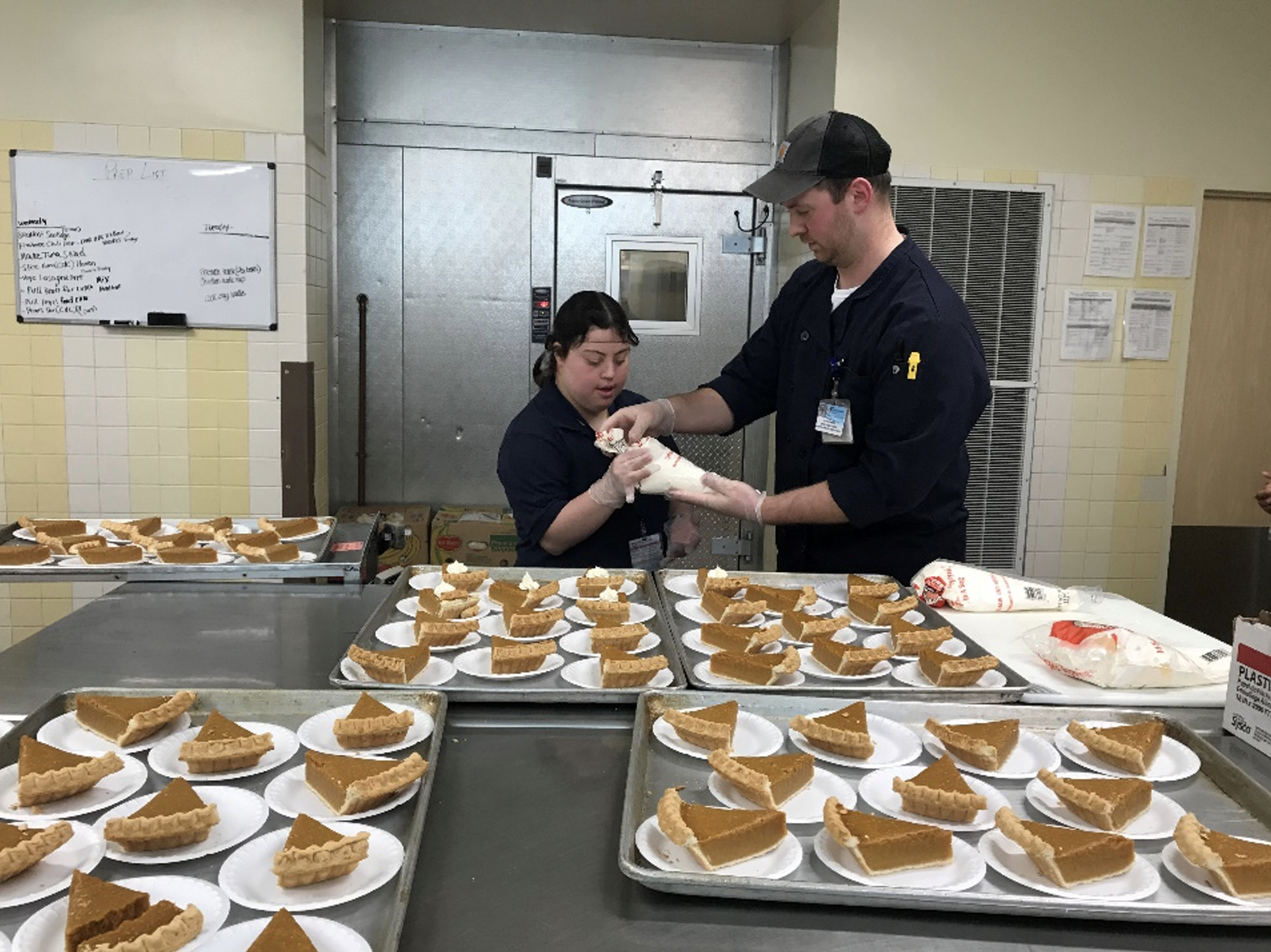A Project SEARCH intern works with a chef in the kitchen as he shows her how to use a piping bag on slices of pumpkin pie.