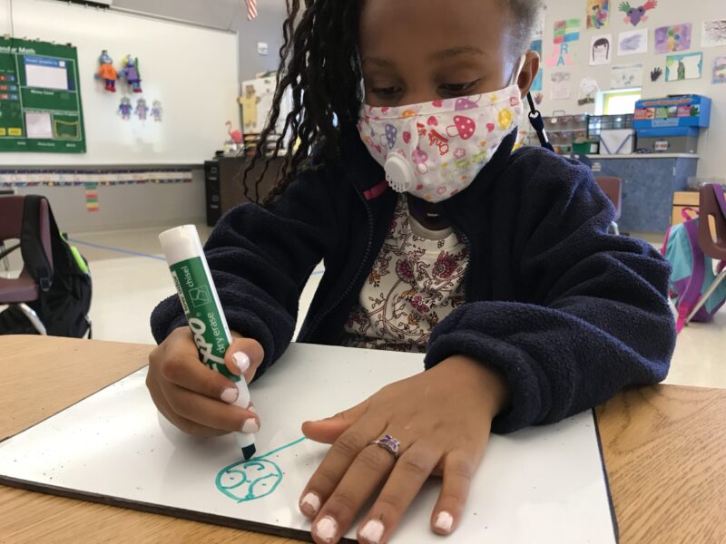 A close-up photo of a kindergarten-aged girl sitting at a desk while drawing in green marker on a tablet-sized white board.