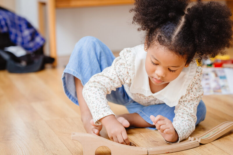 Photo of a young girl playing on the floor with a wooden railway set.