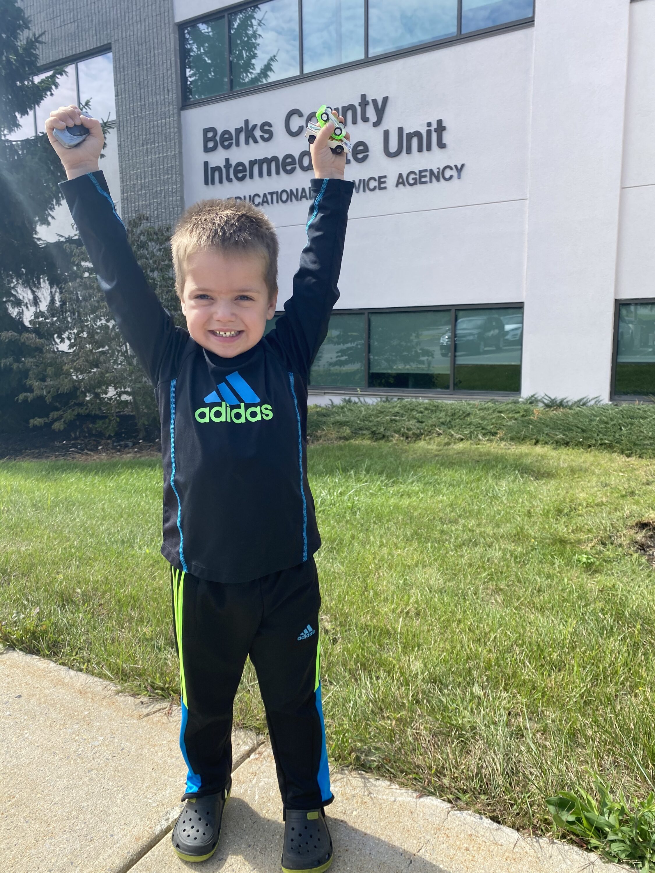 A young boy holding toy cars stands with arms raised in front of a building with the words Berks county Intermediate Unit under the window.